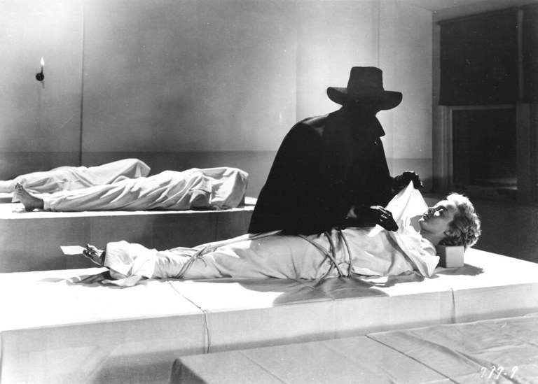 A black and white still from the 1953 film "House of Wax."  Three shrouded bodies lay on cloth-covered tables in a morgue.  It is an austere room with dark, square openings on the wall to the right and a single, gas sconce on the rear wall illuminating the scene.  A figure stands in the center of the frame, visible only as a black sillhouette wearing a wide brimmed hat, gloves, and a long coat with a raised collar.  The figure is pulling the shroud back from the body in the foreground, revealing a white woman's face and blonde curls.
